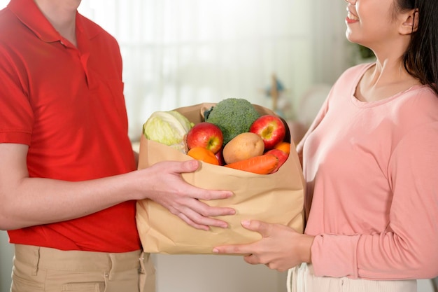 Entrega de um jovem asiático em uniforme vermelho segurando vegetais de frutas saudáveis na sacola de compras