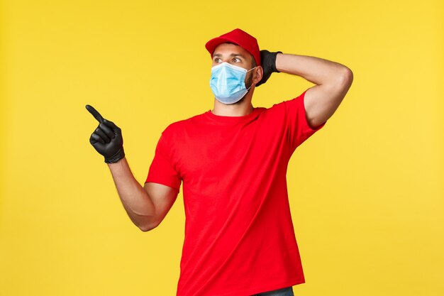 Entrega expressa durante a pandemia, covid-19, frete seguro, conceito de compra online. Correio chocado e impressionado com uniforme vermelho, máscara médica e luvas, apontar e olhar para o canto superior esquerdo