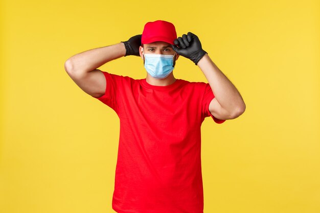 Entrega expressa durante a pandemia, covid-19, frete seguro, conceito de compra online. O mensageiro sério e bonito coloque um boné vermelho uniforme, use máscara e luvas médicas, estando pronto para entregar seu pedido