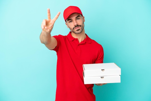 Entrega homem caucasiano pegando caixas de pizza isoladas em fundo azul sorrindo e mostrando sinal de vitória