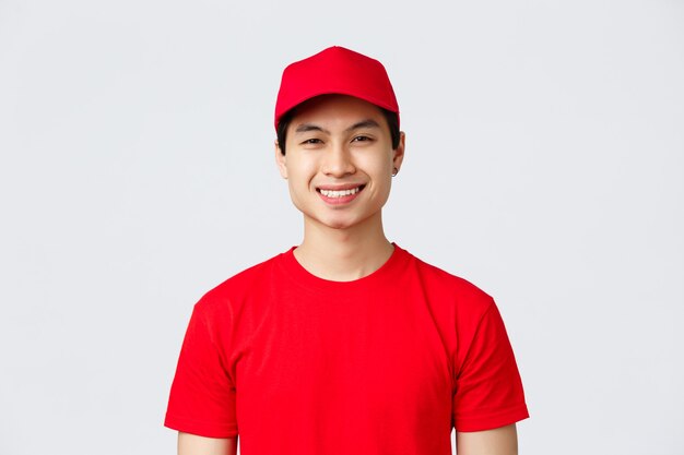 Entrega, pedidos sin contacto y concepto de compra. Sonriente y agradable empleado del servicio de transporte, usa gorra roja uniforme y camiseta, mira la cámara optimista, ayuda al cliente, el mensajero transfiere mercancías.