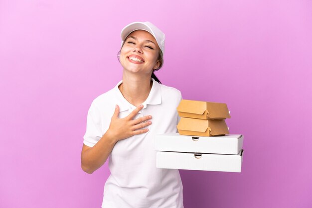 Entrega de pizza Chica rusa con uniforme de trabajo recogiendo cajas de pizza y hamburguesas aisladas en un fondo morado sonriendo mucho