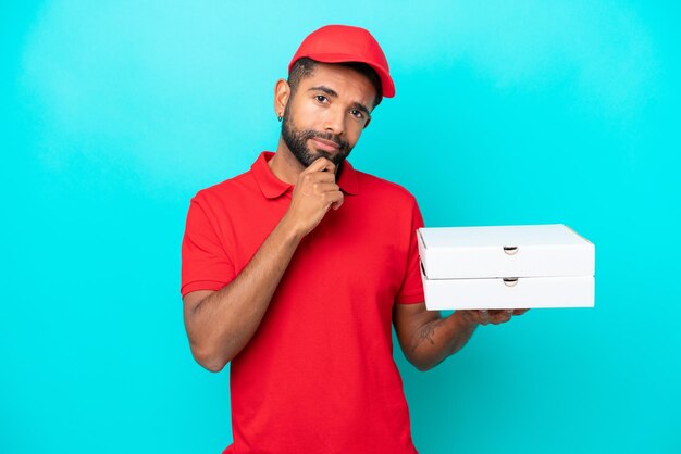 Entrega de pizza Hombre brasileño con uniforme de trabajo recogiendo cajas de pizza aisladas en un pensamiento de fondo azul