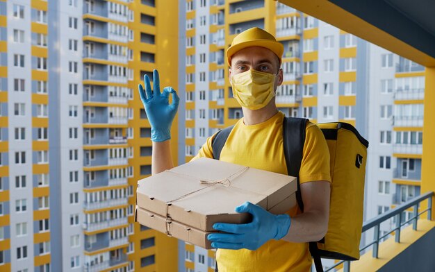 Entrega segura de alimentos. El mensajero con uniforme amarillo, máscara protectora y guantes entrega comida para llevar durante la cuarentena de coronovirus