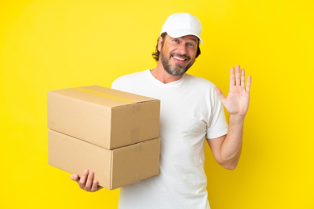 Entrega senior hombre holandés aislado sobre fondo amarillo saludando con la mano con expresión feliz