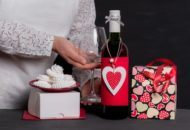 Foto entrega sosteniendo una copa de vino cerca de una botella de vino con un corazón rojo del día de san valentín, una bolsa festiva y pasteles