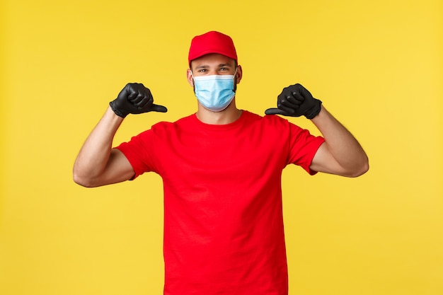 Entrega urgente durante la pandemia, covid-19, envío seguro, concepto de compra. Mensajero sonriente alegre con uniforme rojo y máscara médica, asegura el mejor servicio logístico, confía en él, apuntando a sí mismo.