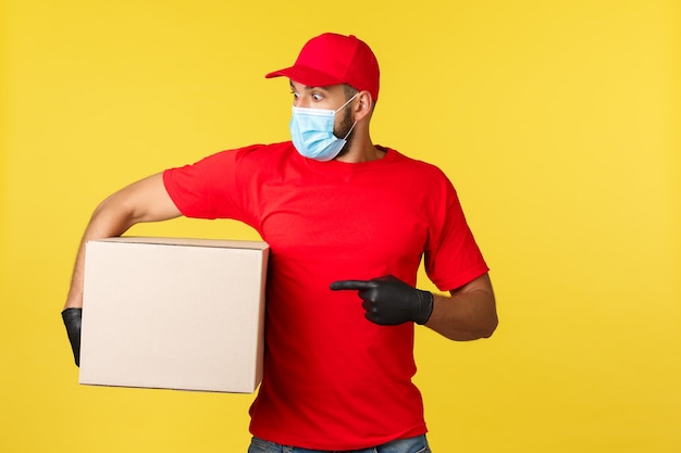 Entrega urgente durante la pandemia, covid-19, envío seguro, concepto de compra online. Mensajero sorprendido y conmocionado en uniforme rojo, máscara médica y guantes, apuntando a la izquierda, sosteniendo el paquete de caja.