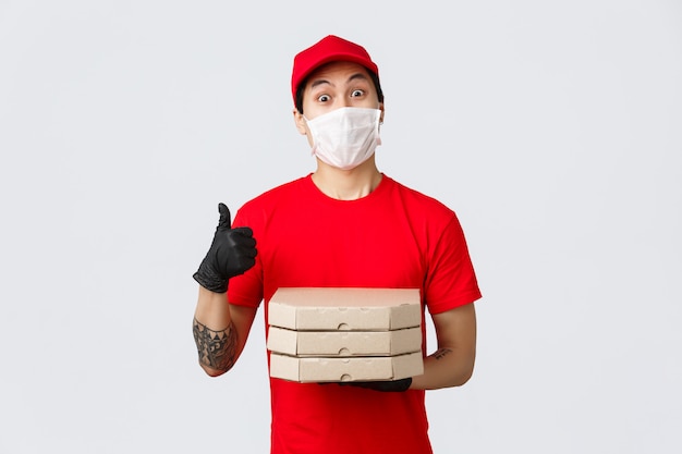 Entregador asiático de uniforme vermelho, boné e camiseta. usando luvas de proteção, máscara médica para a segurança dos clientes durante a entrega de covid-19. segurando caixas de pizza e fazendo o polegar para cima