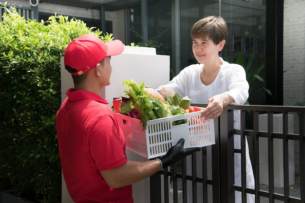 Entregador asiático de uniforme vermelho entregando uma caixa de mantimentos com alimentos, frutas, vegetais e bebidas para uma receptora em casa