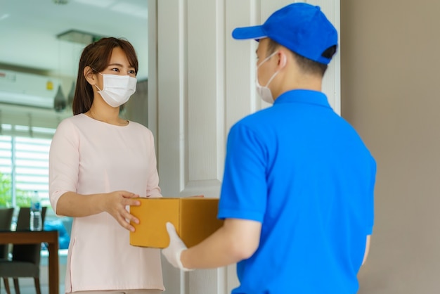 Entregador asiático usando máscara facial e luva de uniforme azul segurando uma caixa de papelão na frente da casa e mulher aceitando a entrega de caixas do entregador durante o surto de COVID-19.