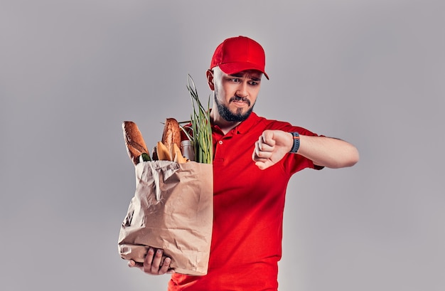 Entregador barbudo chateado jovem com uniforme vermelho segura um pacote com pão e legumes e olha para smartwatch em sua mão atrasando-se isolado em fundo cinza.