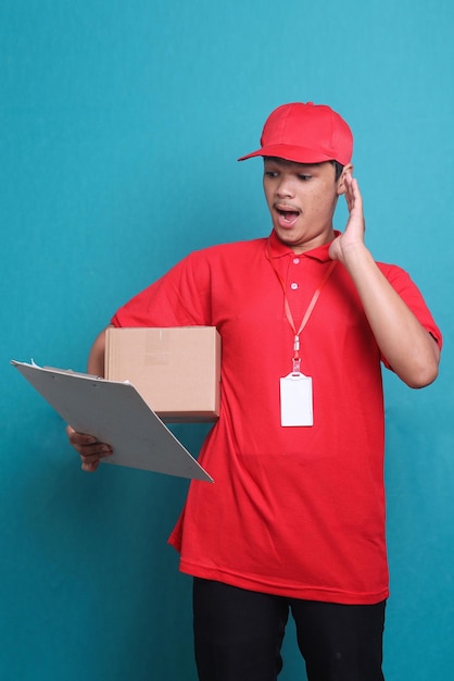 Entregador chocado em uniforme vermelho trabalha como correio de revendedor segura prancheta com caixa isolada em azul