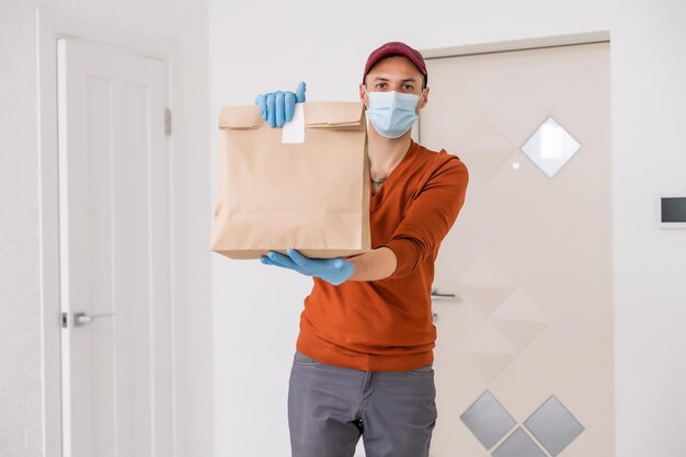 Entregador com máscara protetora e luvas segurando caixa, bolsa com mantimentos em frente a um prédio.