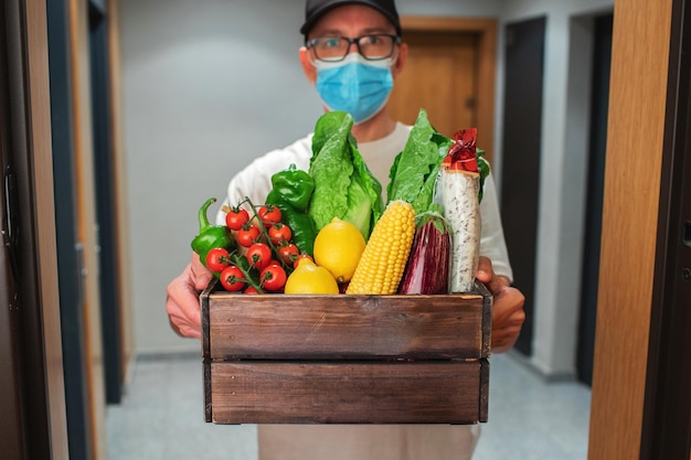 Entregador com máscara protetora segurando o saco de papel com comida na entrada. O mensageiro entrega a caixa com vegetais e frutas frescas ao cliente