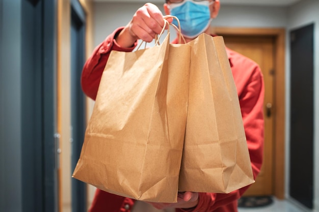 Entregador com máscara protetora segurando o saco de papel com comida na entrada. O mensageiro entrega a caixa com vegetais e frutas frescas ao cliente