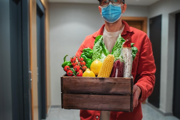 Entregador com máscara protetora segurando saco de papel com comida na entrada O correio entrega a caixa com legumes e frutas frescas ao cliente