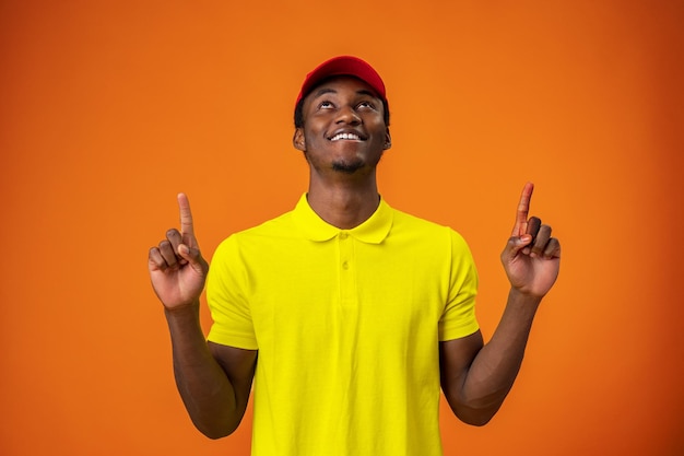 Entregador de camiseta amarela e boné vermelho sorrindo e apontando o dedo para cima contra o fundo laranja