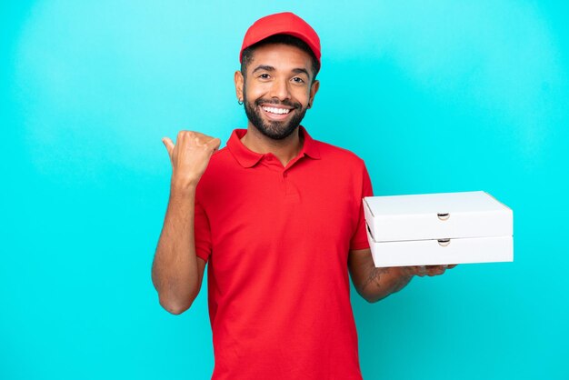 Entregador de pizza brasileiro com uniforme de trabalho pegando caixas de pizza isoladas em fundo azul apontando para o lado para apresentar um produto