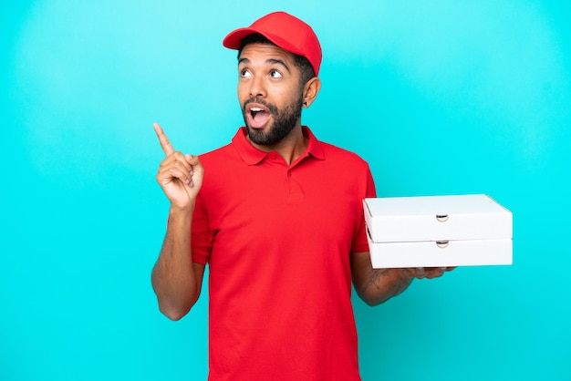 Entregador de pizza brasileiro com uniforme de trabalho pegando caixas de pizza isoladas em fundo azul com a intenção de realizar a solução enquanto levanta um dedo