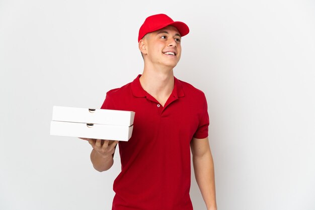 Entregador de pizza com uniforme de trabalho pegando caixas de pizza isoladas na parede branca pensando uma ideia enquanto olha para cima