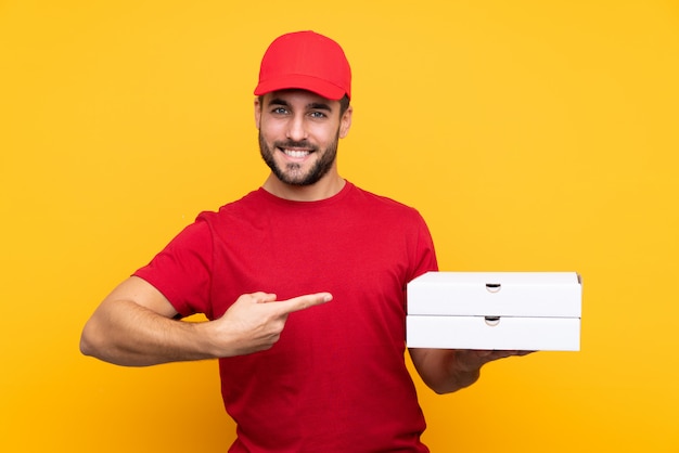 Entregador de pizza com uniforme de trabalho, pegando caixas de pizza sobre parede amarela isolada e apontando-o