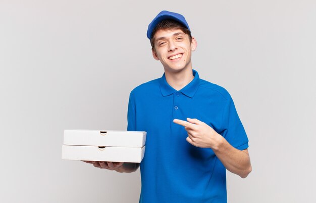 Entregador de pizza jovem sorrindo alegremente, sentindo-se feliz e apontando para o lado e para cima, mostrando o objeto no espaço da cópia