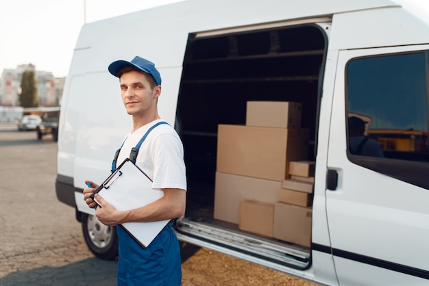 Entregador de uniforme, segurando o pacote e o caderno, caixas de papelão no carro, serviço de entrega. Homem parado diante de pacotes de papelão em um veículo, entregador masculino, correio ou serviço de transporte