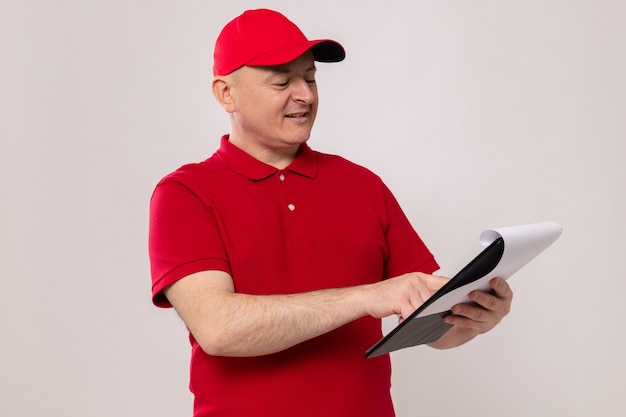 Entregador de uniforme vermelho e boné segurando uma prancheta com páginas em branco olhando para ela com um sorriso no rosto