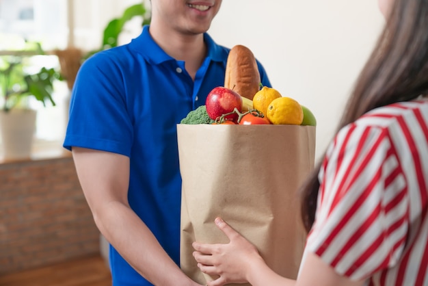 Entregador jovem asiático em uniforme de camisa azul, entregando alimentos frescos de saco de pacote para mulher em casa. Serviço de entrega de mercearia.