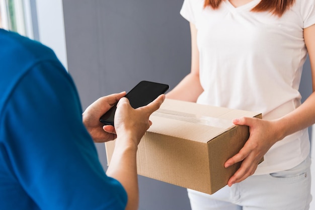 Entregador jovem asiático que faz compras on-line com caixa de correio de pacote em uniforme usando máscara facial protetora fazendo serviço mulher cliente usando assinatura de dedo de mão no telefone móvel digital em casa