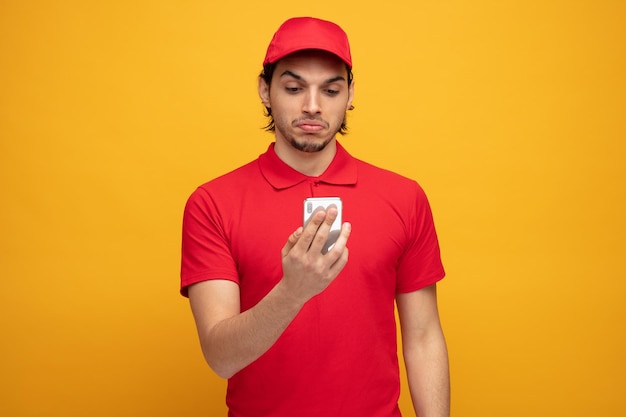 entregador jovem inseguro vestindo uniforme e boné segurando e olhando para o celular isolado em fundo amarelo