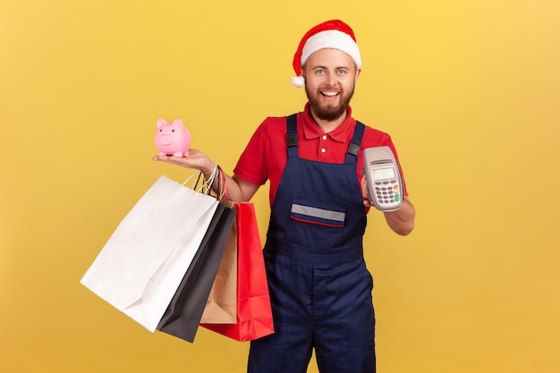 Entregador positivo de uniforme e chapéu de Papai Noel segurando sacolas de papel, terminal de pagamento e cofrinho, pagando sem contato e economizando dinheiro, cashback. Interior isolado em fundo amarelo