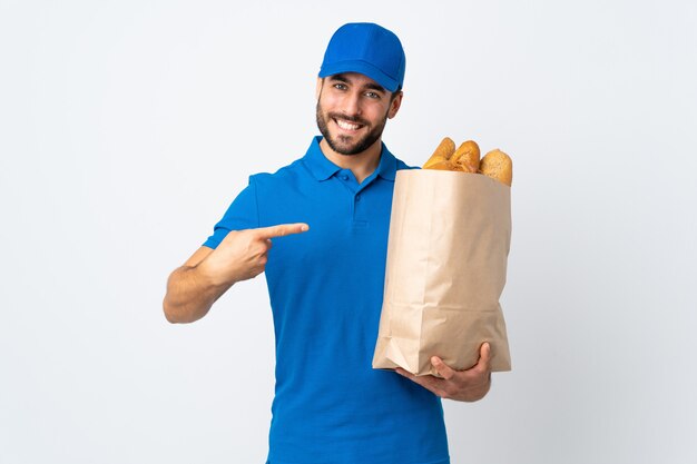 Entregador, segurando uma sacola cheia de pães na parede branca e apontando-o