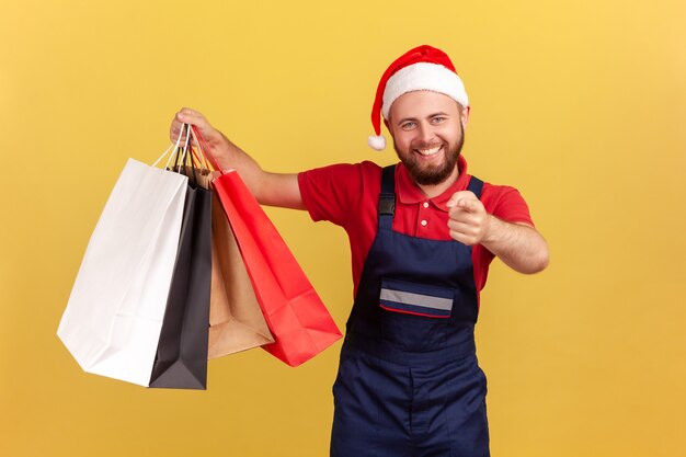 Entregador sorridente positivo com chapéu de papai noel segurando sacolas de papel e apontando o dedo para você,