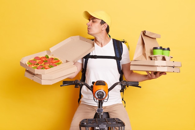 Entregador vestindo boné e camiseta branca segurando caixas de pizza e café andando de bicicleta cheirando delicioso pedido com os olhos fechados e curtindo posar isolado no fundo amarelo