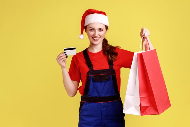 Entregadora de boa aparência mostrando cartão de crédito e sacolas de compras transporte rápido para férias de inverno vestindo macacão azul e chapéu de papai noel Tiro de estúdio interno isolado em fundo amarelo