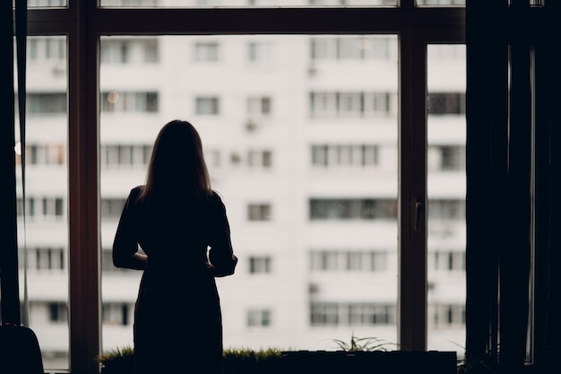 Entrenador adulto joven de mujer de negocios de pie en la silueta de la ventana de la oficina