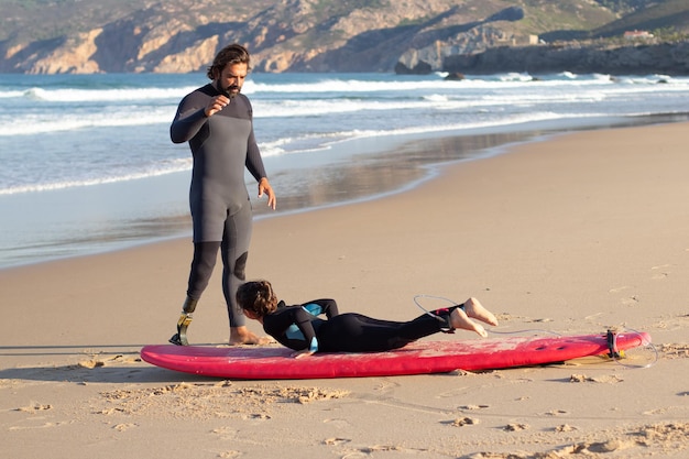 Entrenador atento con pierna mecánica y chica cerca del océano. Hombre adulto medio enseñando a una niña de cabello oscuro cómo usar tablas de surf. Clase, ocio, concepto de estilo de vida activo.