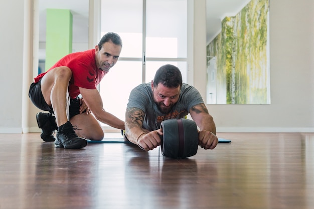 Entrenador ayudando a un hombre a hacer ejercicio con un rodillo de abdominales en un gimnasio