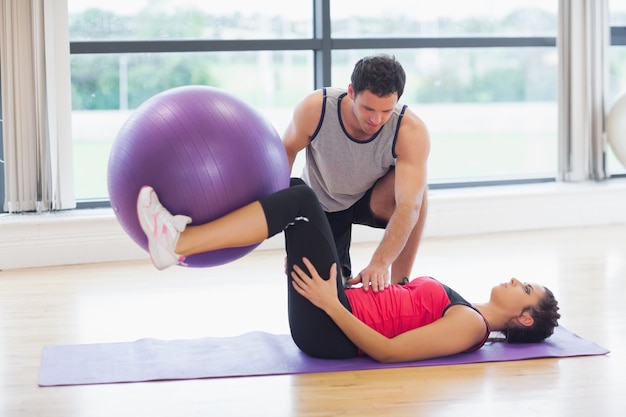 Entrenador ayudando a la mujer joven con la bola de la aptitud en el gimnasio