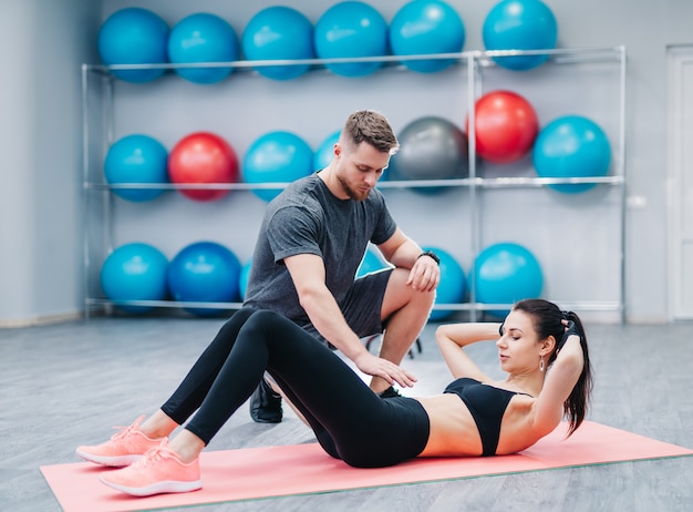 Entrenador ayudando a una mujer joven a hacer ejercicios abdominales en el fondo de pelotas de fitness