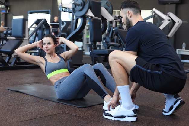 Entrenador ayudando a la mujer joven a hacer ejercicios abdominales en el gimnasio.