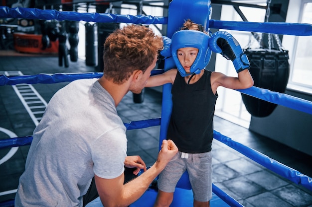 Entrenador de boxeo joven está ayudando a niño en ropa protectora en el ring entre rondas.