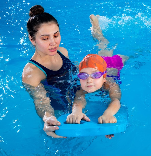 Entrenador enseñando a una niña a nadar en una piscina cubierta con una tabla flotante en la piscina