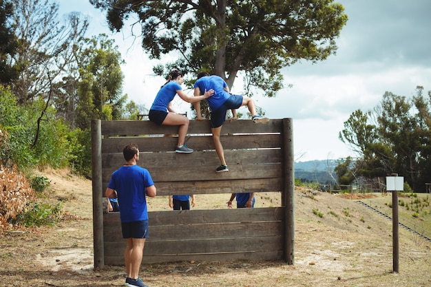 Entrenador femenino ayudando al hombre a trepar por encima de la pared de madera durante la carrera de obstáculos