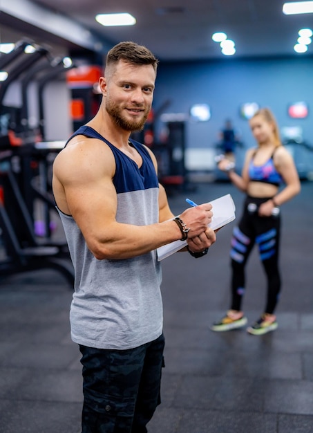 Entrenador de fitness personal con portapapeles. Chica fitness agradable con figura delgada sobre fondo borroso. Fondo de gimnasio moderno.