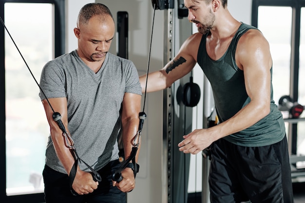 Entrenador de fitness que controla la postura del cliente que hace ejercicio en la máquina de cable cruzado para fortalecer los músculos de los brazos y la parte superior del pecho
