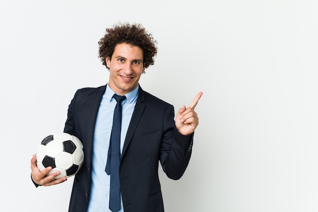 Entrenador de fútbol sosteniendo una pelota sonriendo alegremente señalando con el dedo lejos.