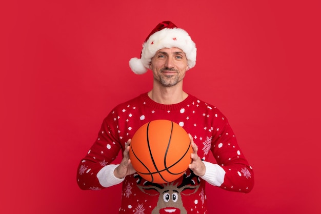 Entrenador de hombre sostenga la pelota de baloncesto en suéter de vacaciones y gorro de Papá Noel en el fondo del estudio Venta de Navidad u oferta especial de invierno